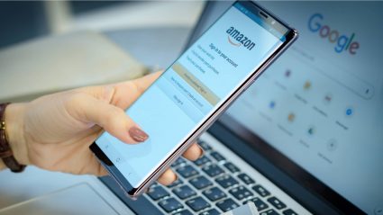 Person using mobile phone with Amazon website open, with laptop in background displaying Google.com