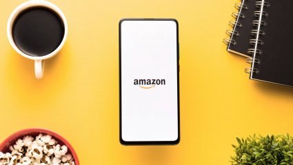Amazon app displayed on a mobile phone, atop a table next to popcorn, coffee, a houseplant, and two notebooks