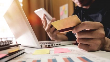 Man sitting in front of laptop, holding mobile phone in one hand and credit card in the other