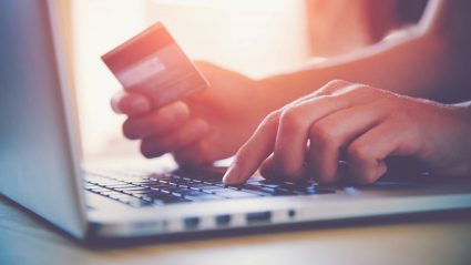 A person typing on a laptop with one hand and holding a credit card in the other hand