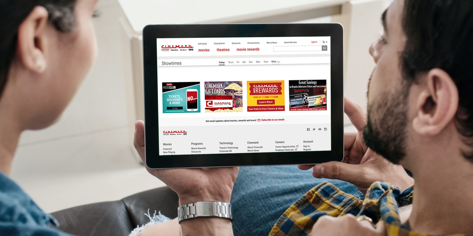 Two people holding a tablet displaying a website for movie showtimes, rewards, and ticket purchasing options.