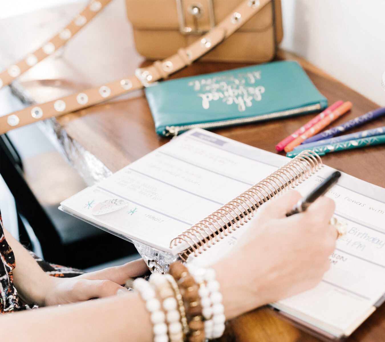 A person writes in a spiral planner on a wooden desk with pens, a beaded bracelet, a purse, and a teal pouch nearby.