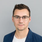 A young man with short brown hair, glasses, and slight facial hair, wearing a navy blazer and a light-colored shirt, poses in front of a plain gray background.