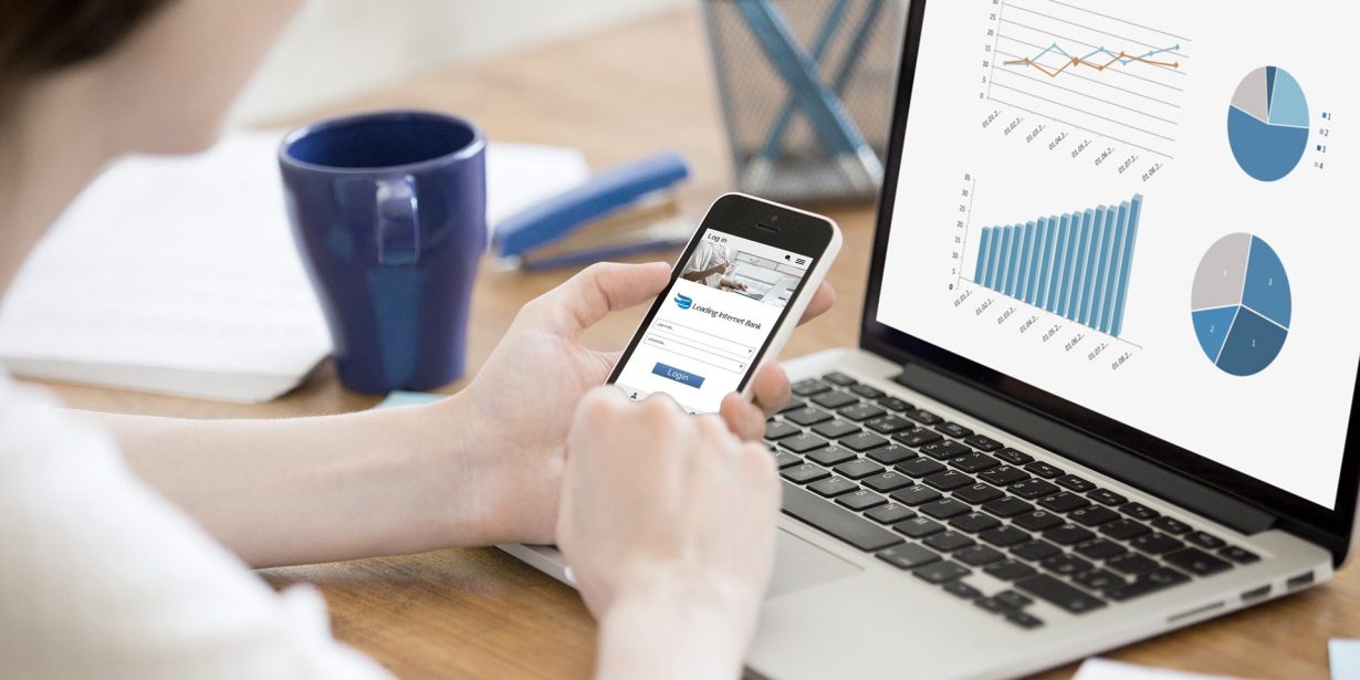 Person using smartphone and laptop at desk; laptop screen displays line and bar graphs, and pie charts; blue mug and papers in background.