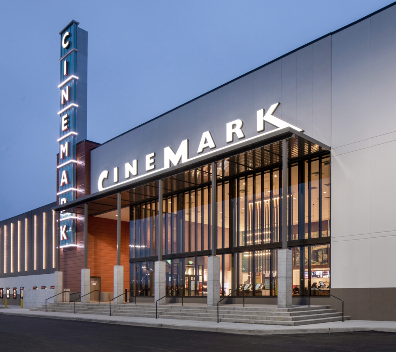 Exterior view of a modern Cinemark movie theater with large glass windows, illuminated signs, and a wide entrance with stairs.