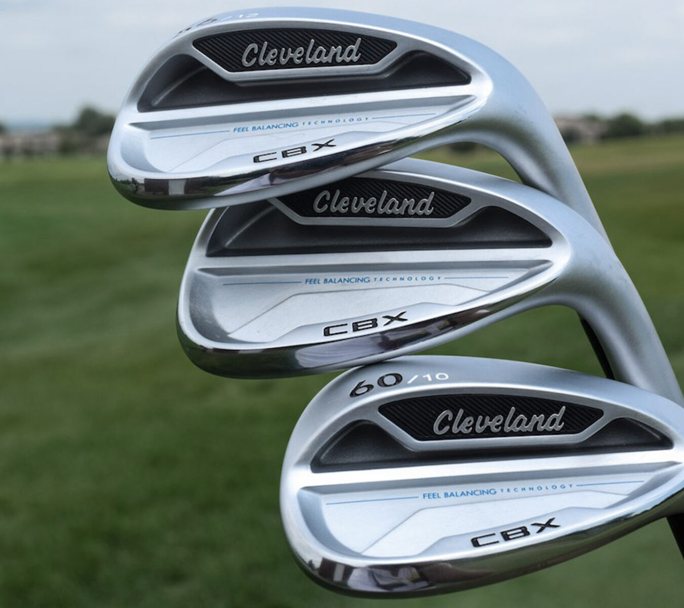 Three Cleveland CBX golf irons with “Feel Balancing Technology” are stacked together outdoors on a grassy area.