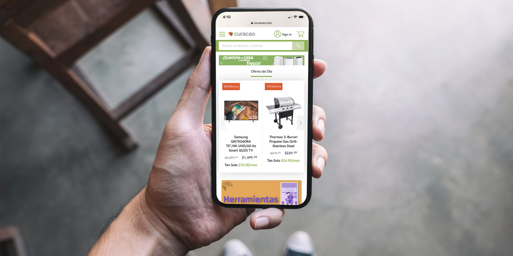 A person holds a smartphone displaying an online shopping app with product listings for a Samsung TV and a Char‑Broil grill.
