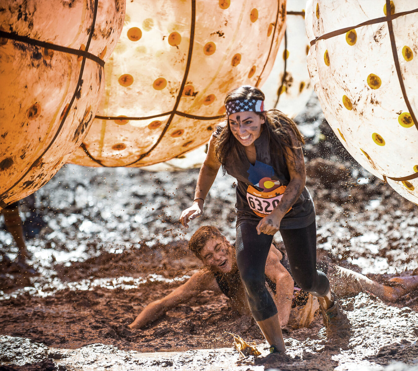 A woman runs through a muddy obstacle course, smiling, while a man falls behind her; large inflatable balls hang above them.
