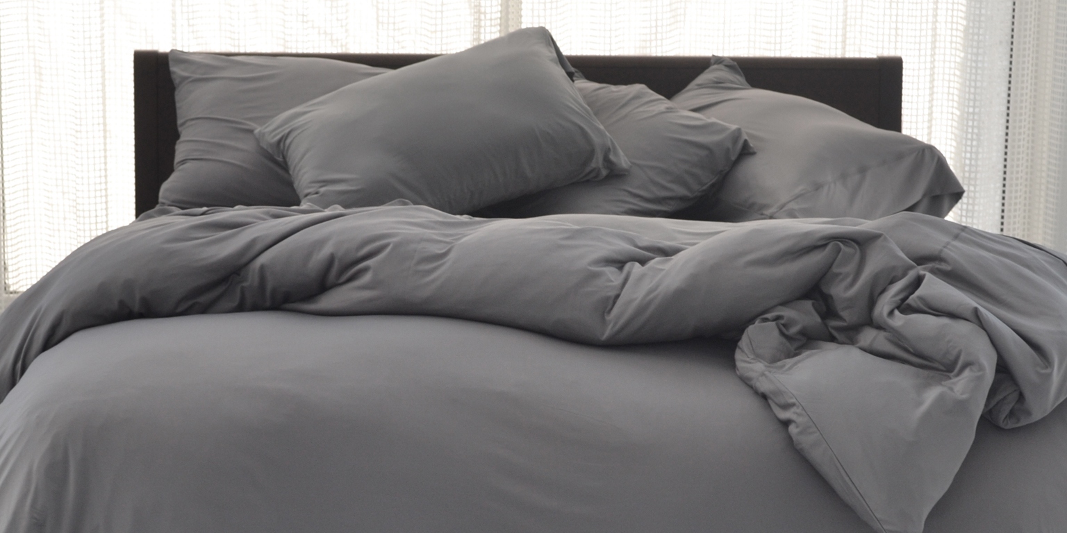 A neatly made bed with gray bedding, including pillows and a comforter, in front of sheer white curtains.