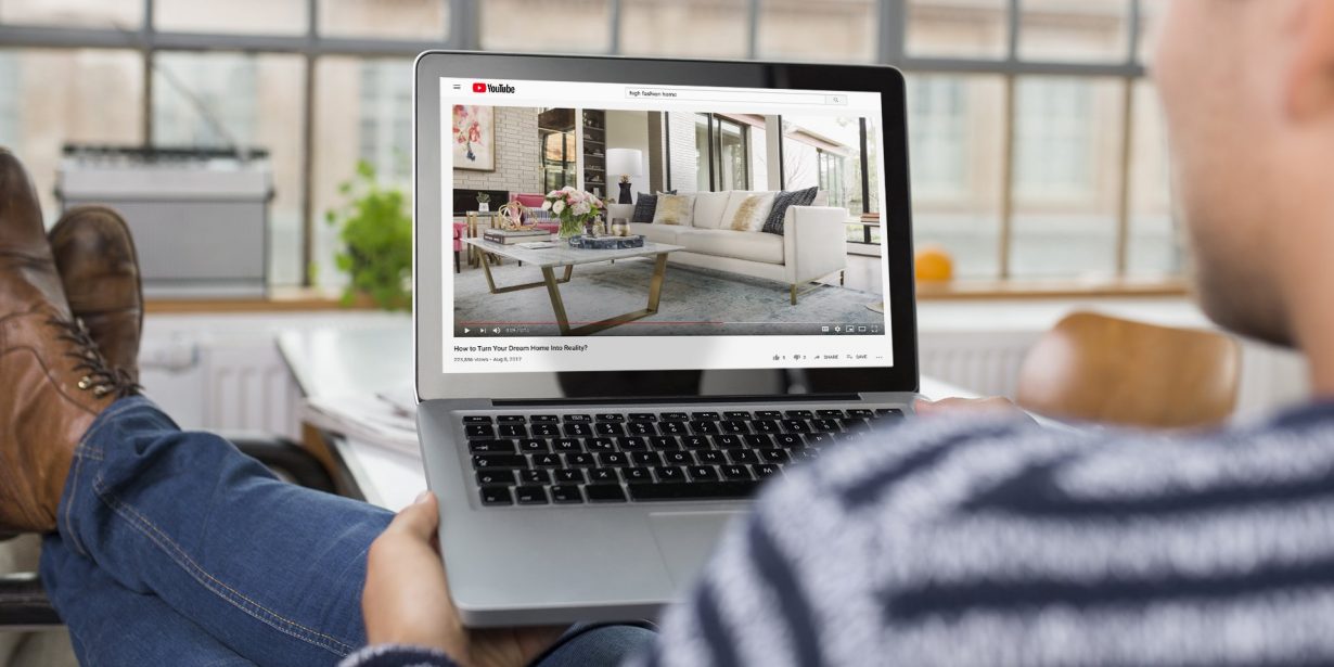 Person sitting with a laptop watching a YouTube video featuring a modern living room interior with a white sofa and glass coffee table.