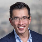 A man wearing glasses and a navy blue suit jacket smiles at the camera against a blurred background.