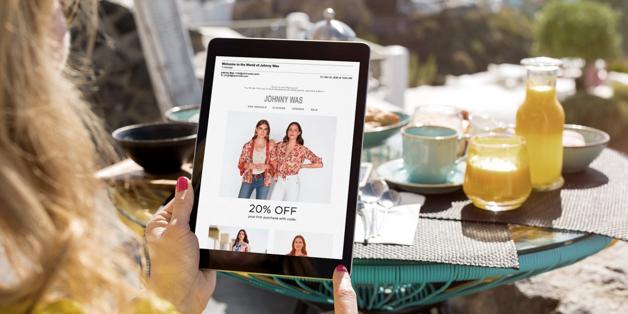 Person holding a tablet with a Johnny Was online store page showing a 20% off sale, seated at an outdoor table with breakfast items including juice, coffee, and cups.