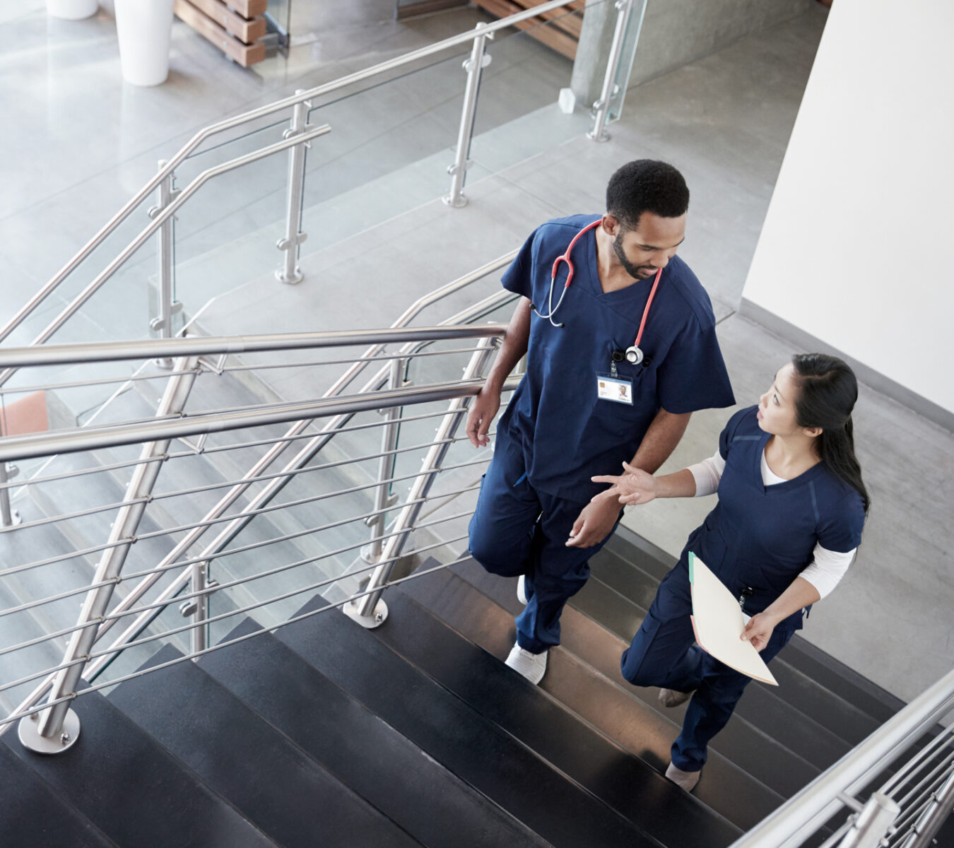 Two healthcare professionals in scrubs walk up a staircase indoors, conversing. One carries a folder and both wear stethoscopes.