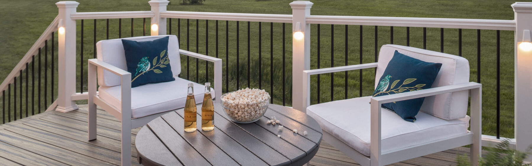 Two white patio chairs with blue leaf-patterned pillows and a round table holding popcorn and two bottled drinks on a deck with a grassy lawn in the background.