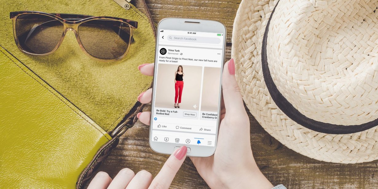 A person holding a smartphone displaying a Facebook post, with sunglasses, a yellow bag, and a straw hat on a wooden surface in the background.