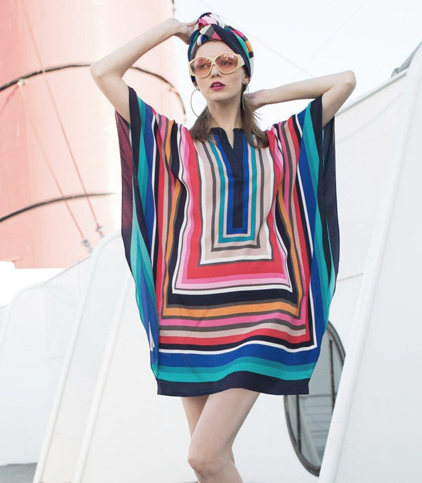 A woman wearing a colorful, geometric-patterned dress, sunglasses, and a matching headscarf poses outdoors against a white and red modern structure.