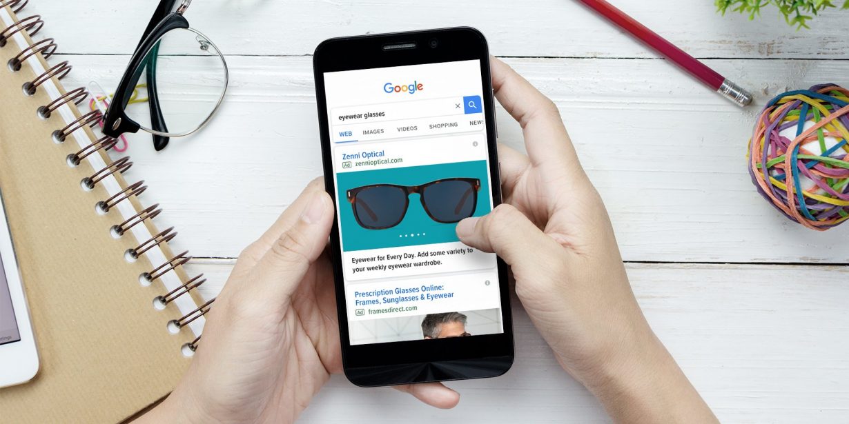 A person holding a smartphone searches for "eyewear glasses" on Google at a desk with a notebook, pen, paper clips, and a ball of rubber bands.