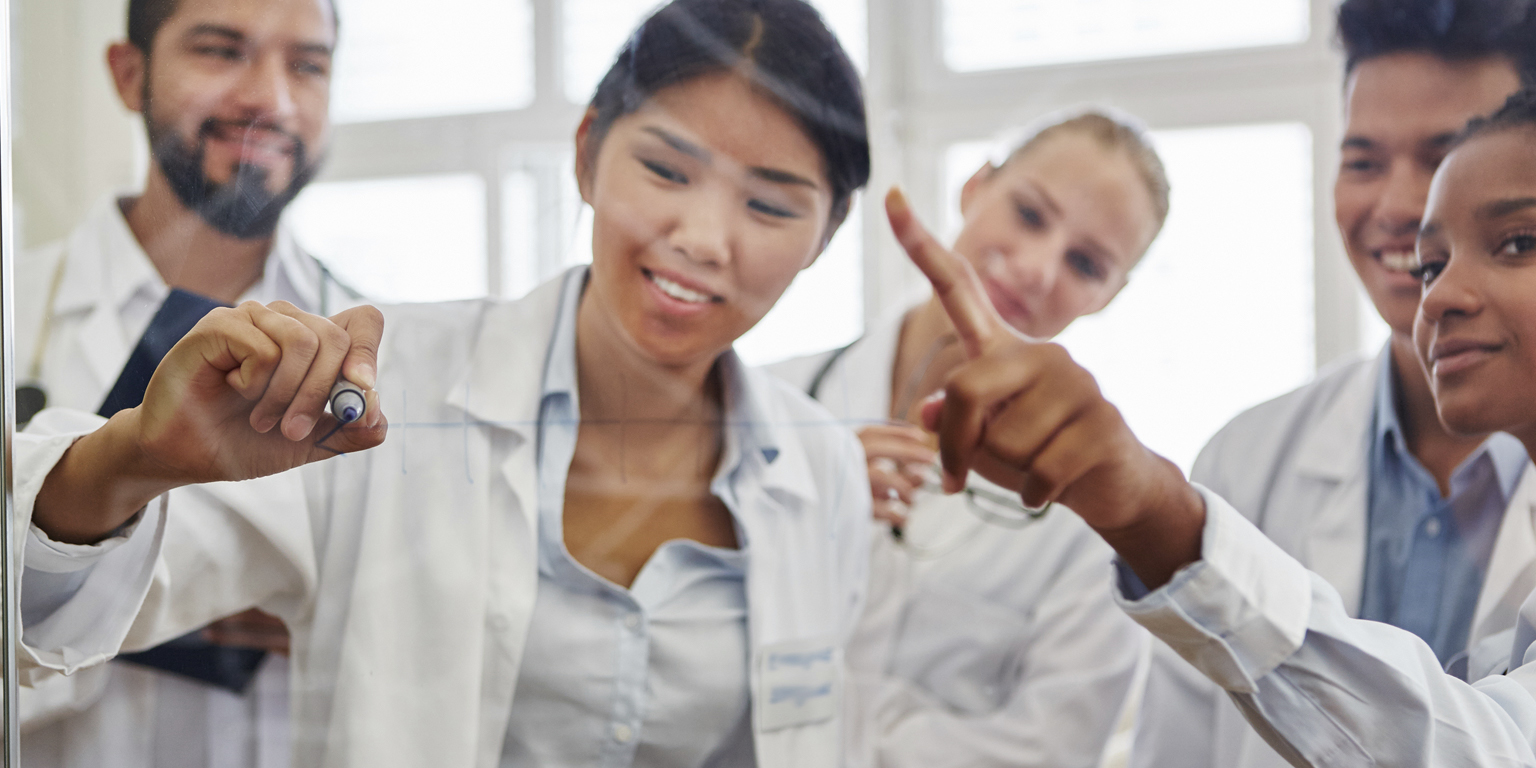 A group of people in lab coats collaborate, with one person drawing or writing on a transparent surface while others observe and discuss.