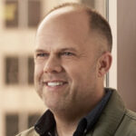 A person with short hair and a beard, wearing a green jacket and dark shirt, smiles while standing indoors near a window.