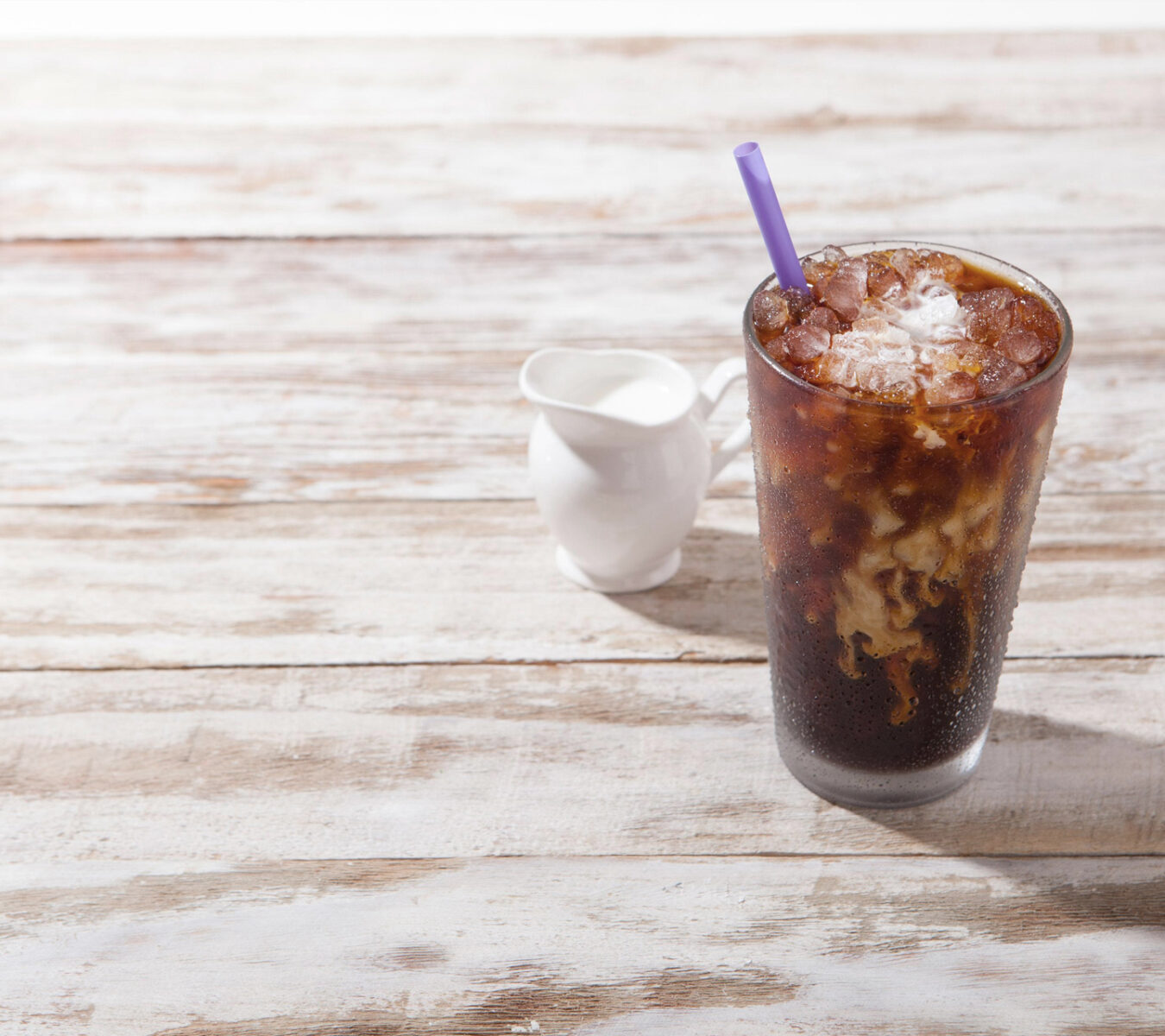 A glass of iced coffee with a purple straw sits beside a small white creamer on a rustic wooden table.