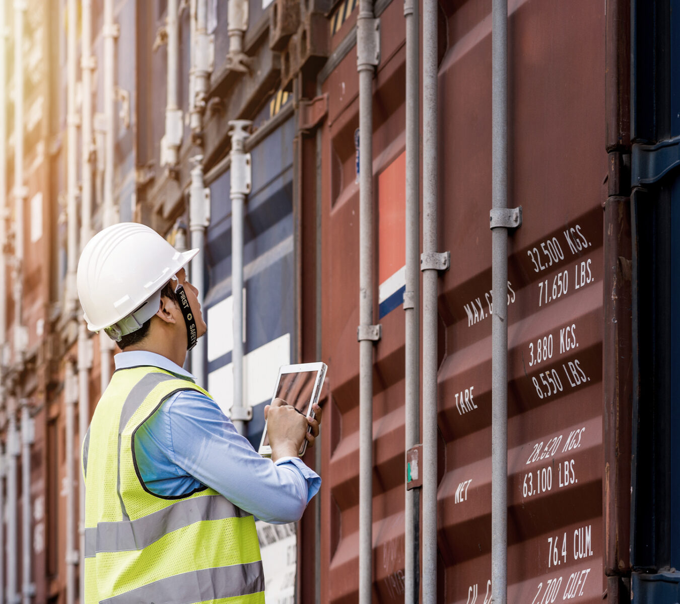A worker in a hard hat and safety vest uses a tablet to inspect stacked shipping containers with visible weight markings.