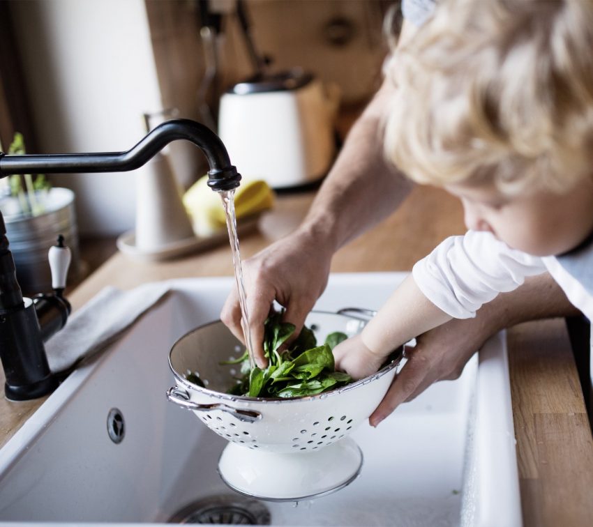 Adult and child washing fresh spinach leaves in a colander under running water at a kitchen sink.