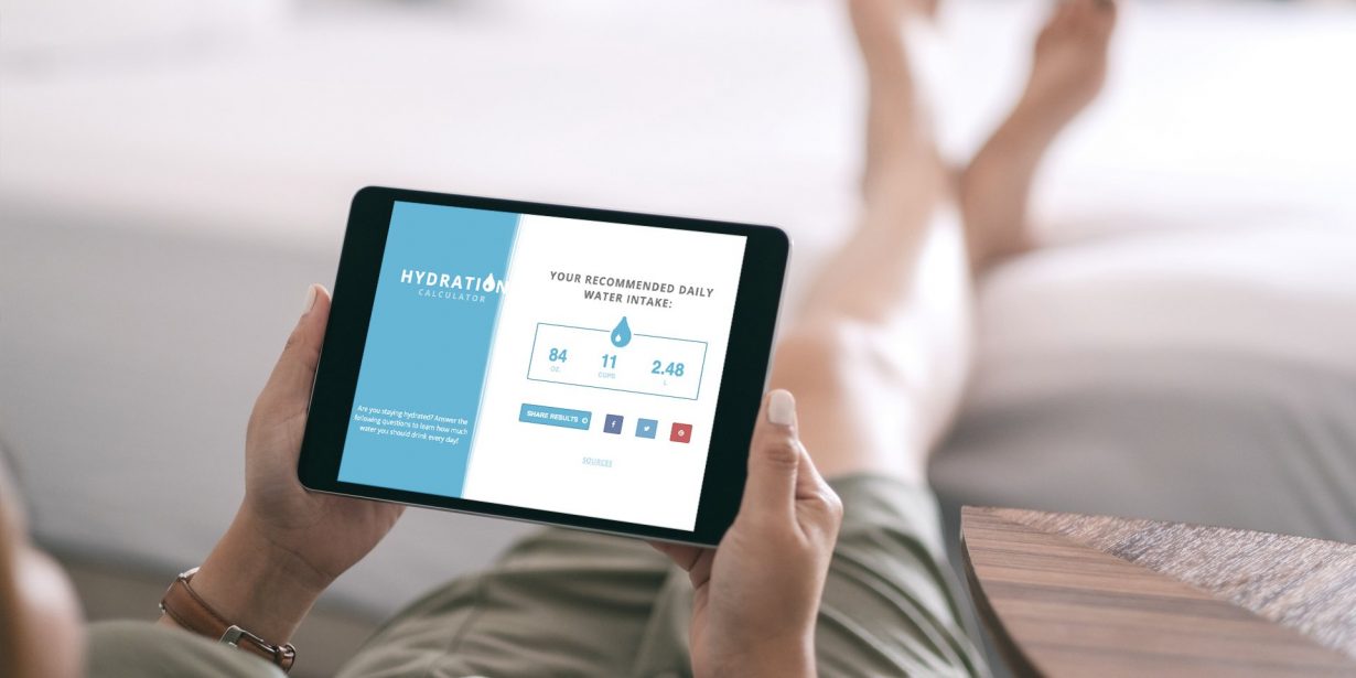 Person holding a tablet showing a hydration calculator with water intake recommendations and metrics, while sitting with legs extended on a light-colored surface.