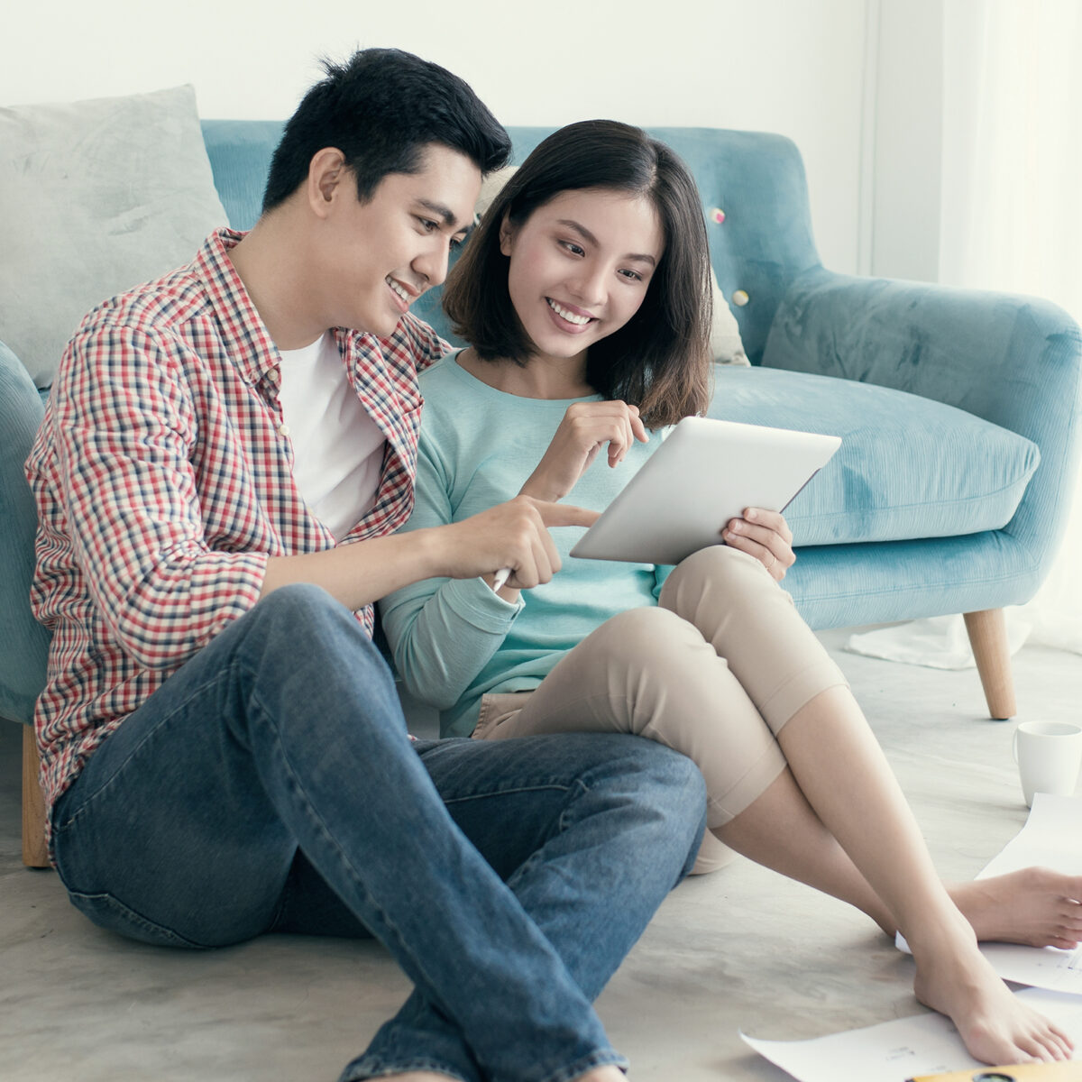 A man and woman sit on the floor in front of a sofa, smiling and looking at a tablet together.
