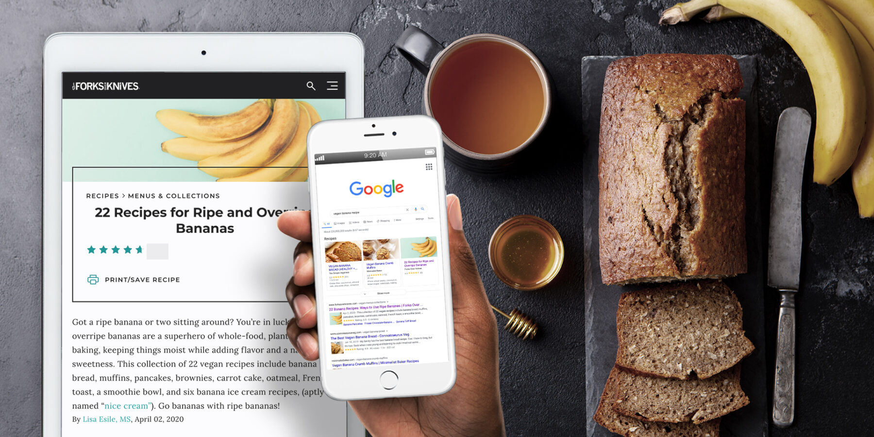 A sliced banana bread loaf on a cutting board with bananas, a cup of tea, and a person holding a phone and a tablet showing banana recipes.