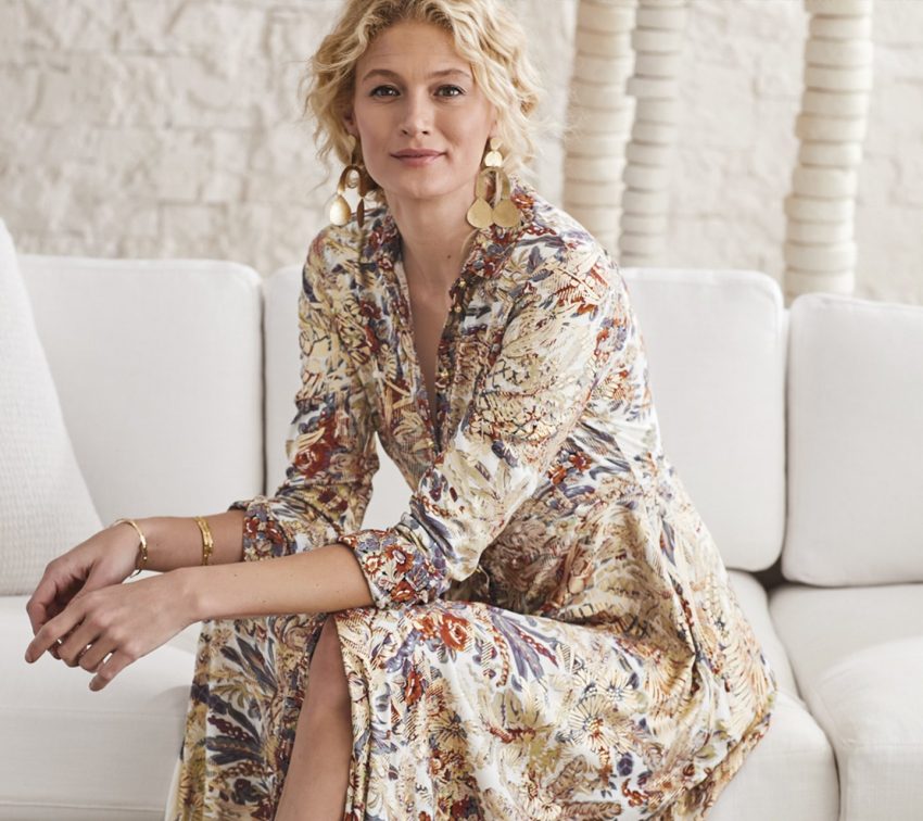 Woman with curly blonde hair sits on a white couch, wearing a long, floral-patterned dress and large round earrings, with a neutral background behind her.