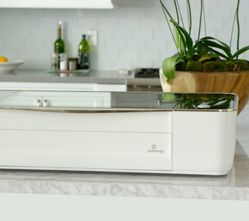 A white Glowforge laser cutter sits on a marble kitchen counter with a wooden planter of green plants in the background.