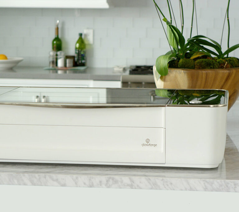 A white Glowforge laser cutter sits on a marble kitchen counter with a wooden planter of green plants in the background.