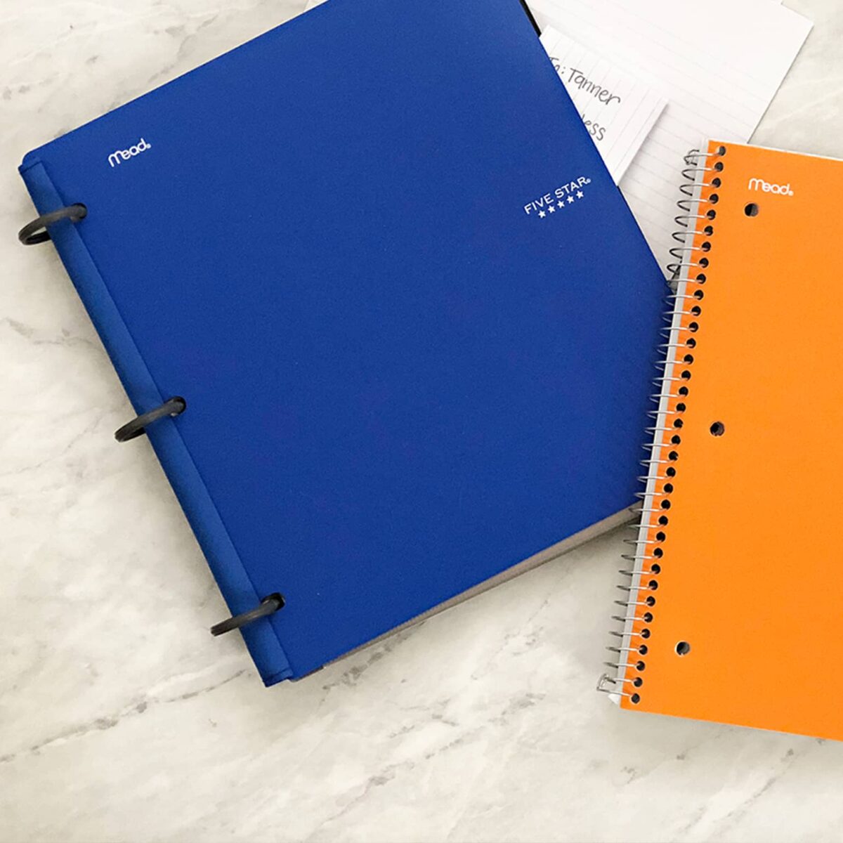 Two Mead notebooks, one blue and one orange, sit on a white marble surface next to a stack of index cards with handwriting partially visible.