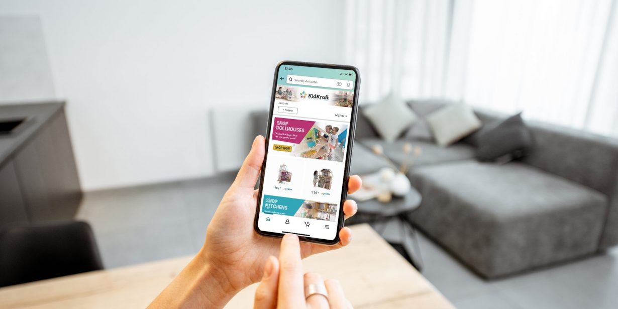 Person holding a smartphone browsing an online clothing store in a modern living room with a gray sofa and large windows in the background.