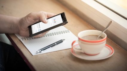 A person sitting by a window with a notepad and cup of coffee, viewing Google on their mobile phone