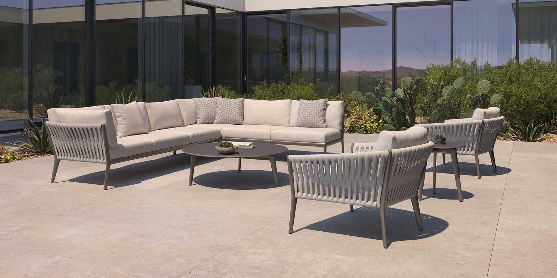 Modern outdoor patio set with a beige sectional sofa, two matching armchairs, and a round black coffee table on a concrete patio, surrounded by desert plants and a glass-walled building.