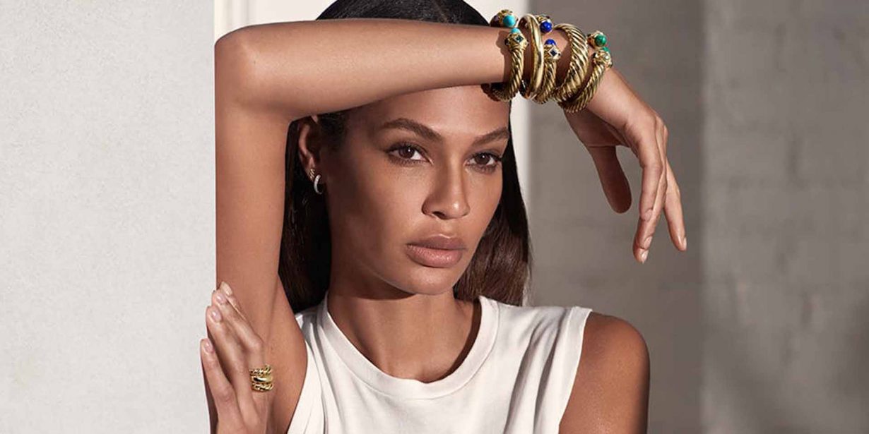 A woman with long hair poses indoors, resting her arm on her forehead, wearing a white sleeveless top and multiple gold bracelets and rings.