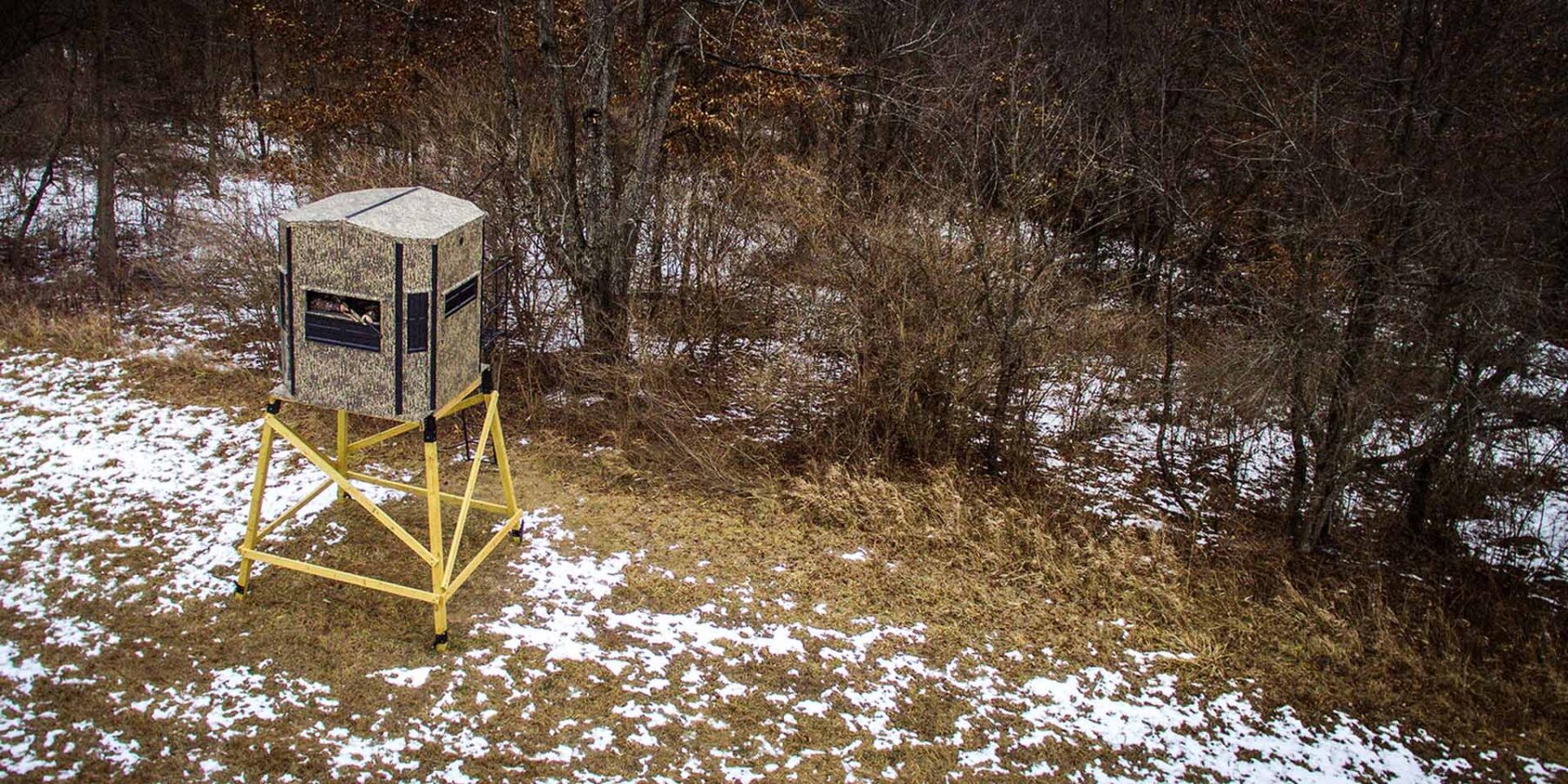 A camouflage hunting blind elevated on wooden stilts stands near a tree line, with sparse snow covering the ground and brown grass visible.