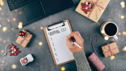 Person writing a holiday shopping list on a clipboard surrounded by presents and gift wrap