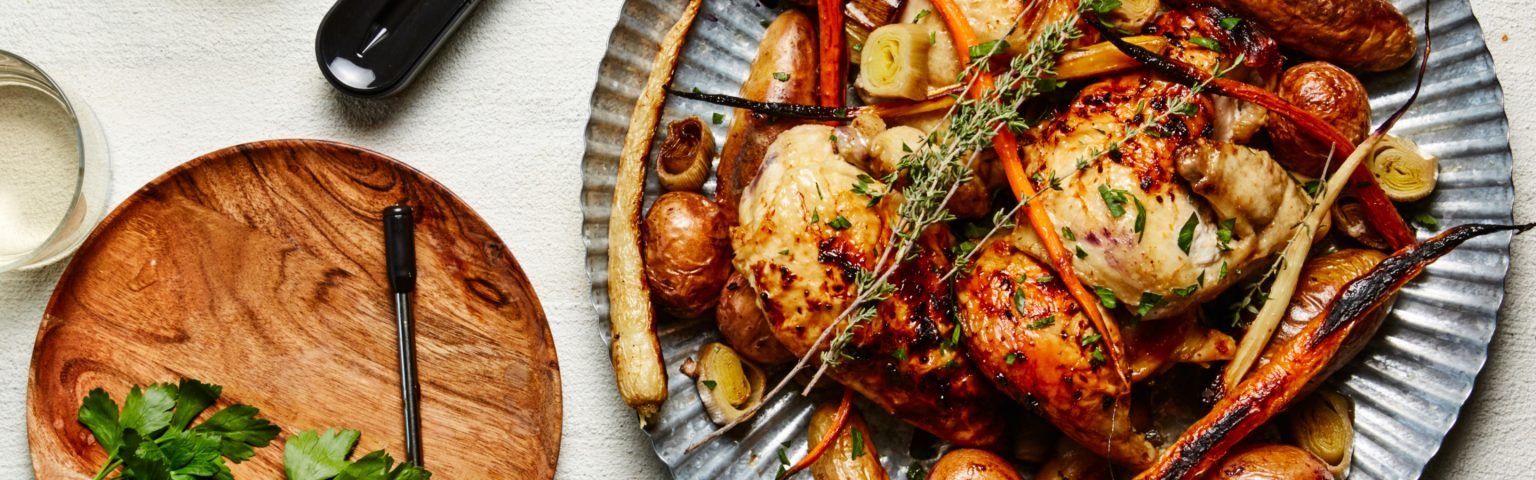 Roast chicken with herbs, carrots, potatoes, and parsnips on a metal platter, next to an empty wooden plate and a glass of water.