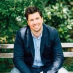 A man in a navy blazer and light blue shirt sits on a wooden bench outdoors, smiling at the camera with greenery in the background.