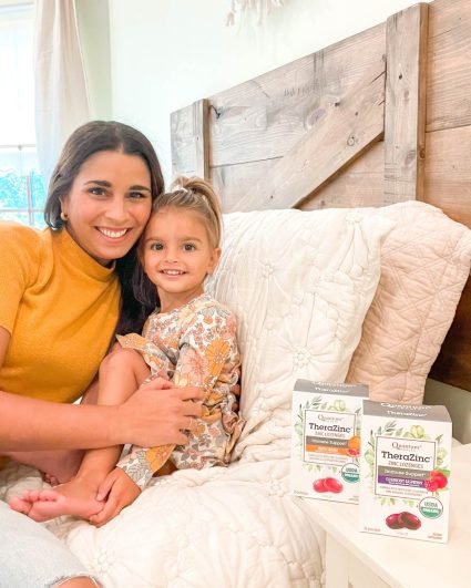 A mother and daughter sitting on a bed beside two boxes of Quantum Health zinc lozenges on a bedside table