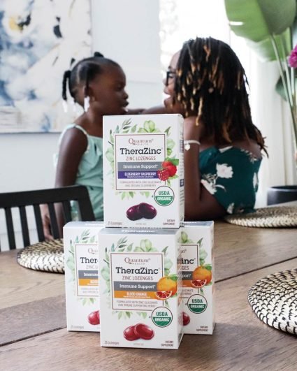 Four boxes of Quantum Health zinc lozenges stacked on a table in the foreground with a mother and daughter in the background