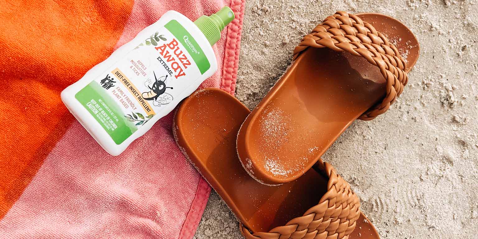 Quantum Health Buzz Away bug spray on a beach blanket beside a pair of sandals