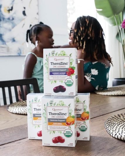 Three boxes of Quantum Health TheraZinc lozenges are on a table, with a woman and child sitting together in the background.