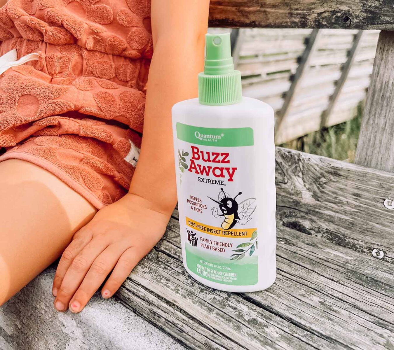 A child sits on a wooden bench next to a bottle of Buzz Away Extreme insect repellent spray.