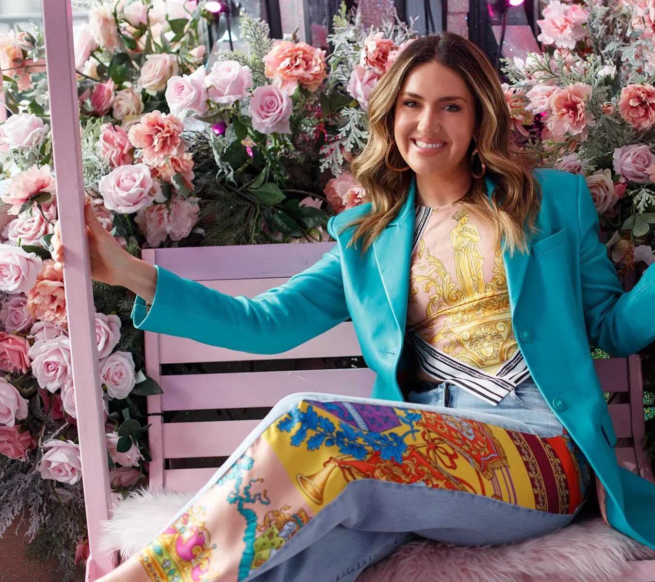 Woman sits on a pink swing decorated with pink roses, wearing a turquoise blazer and colorful patterned pants, smiling at the camera.