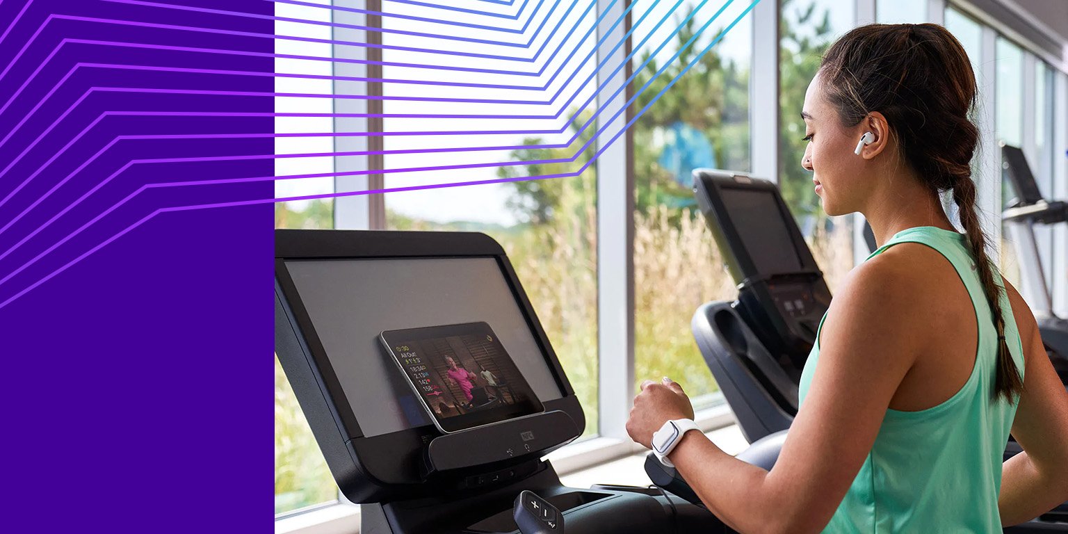 Woman in athletic wear using a treadmill while watching a screen, with large windows and natural light in the background.