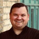 A man with short dark hair and a beard smiles at the camera while standing in front of a brick wall and teal door.
