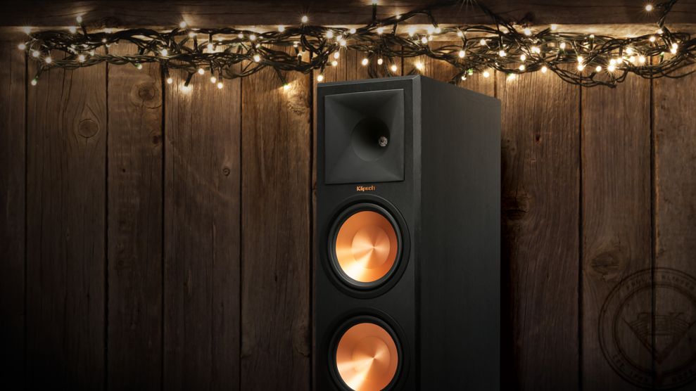 A black Klipsch speaker with copper cones is placed in front of a wooden wall decorated with a string of white fairy lights.